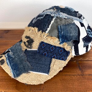 Op de afbeelding: Een bruine, beige en blauwe patchwork baseballcap met een rafelige rand. De cap is gemaakt van verschillende stoffen, waaronder denim, jute en canvas.