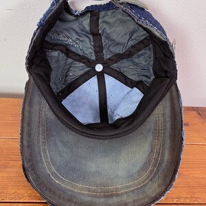 May include: A dark blue denim baseball cap shown from the interior perspective, featuring a frayed edge brim, black interior lining tape, a central button, and an adjustable strap with a metal buckle closure at the back of the head opening.