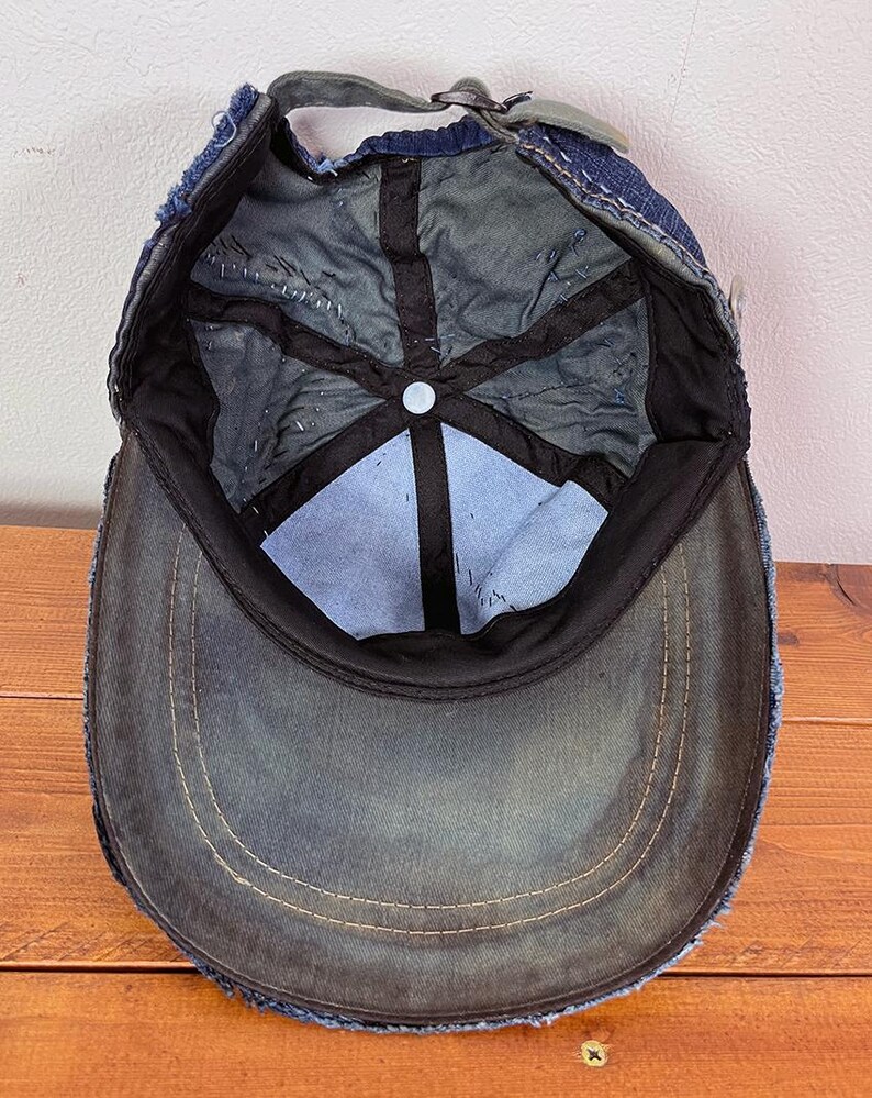 May include: A dark blue denim baseball cap shown from the interior perspective, featuring a frayed edge brim, black interior lining tape, a central button, and an adjustable strap with a metal buckle closure at the back of the head opening.