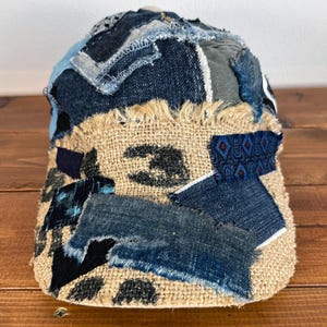 Op de afbeelding: Een bruine en beige patchwork-pet gemaakt van denim en jute. De pet heeft een rafelige rand en is versierd met verschillende patches in verschillende kleuren en texturen.
