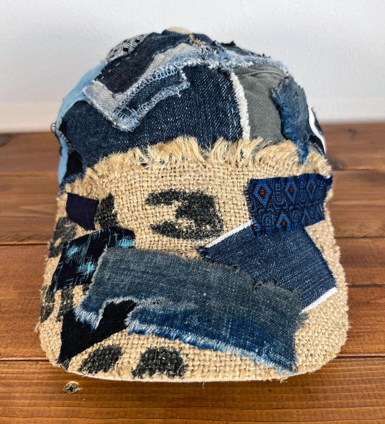 Op de afbeelding: Een bruine en beige patchwork-pet gemaakt van denim en jute. De pet heeft een rafelige rand en is versierd met verschillende patches in verschillende kleuren en texturen.