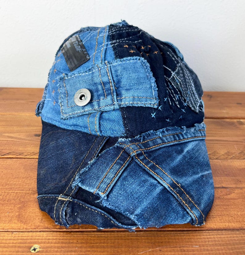 Op de afbeelding: Een blauwe denim baseballcap gemaakt van patchwork denim stof. De cap heeft verschillende stikdetails en een knoop bovenop.