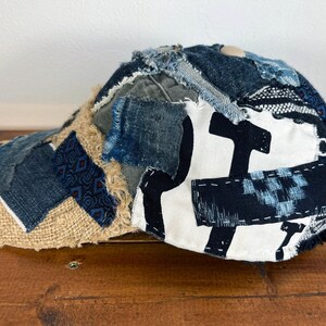 Op de afbeelding: Een blauwe en bruine patchwork baseballcap met een witte canvas kroon met een zwart grafisch ontwerp. De cap is gemaakt van verschillende denim en stoffen resten.