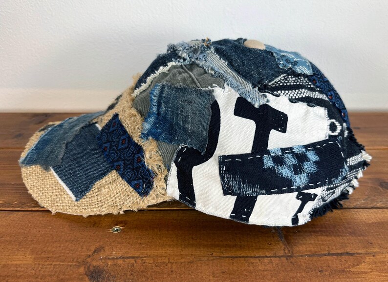 Op de afbeelding: Een blauwe en bruine patchwork baseballcap met een witte canvas kroon met een zwart grafisch ontwerp. De cap is gemaakt van verschillende denim en stoffen resten.