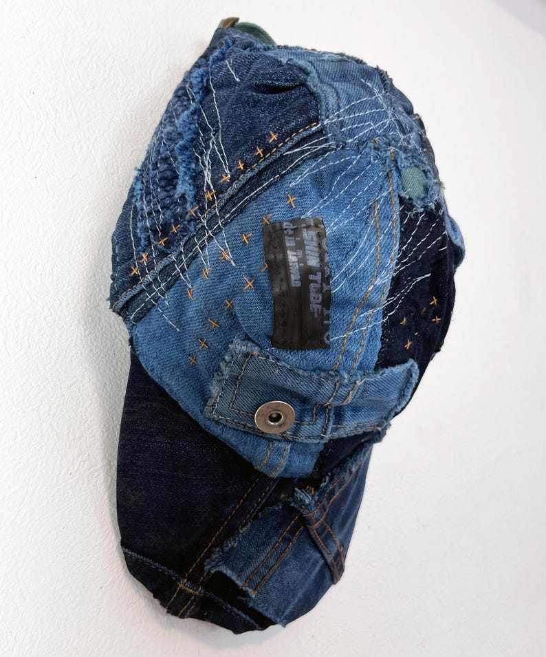 Op de afbeelding: Een blauwe denim baseballcap met een patchwork-ontwerp. De cap is gemaakt van verschillende stukken denim die aan elkaar zijn genaaid, en heeft een rafelige rand. De cap heeft een klein zwart label met de tekst "SKIN TUBE" erop.