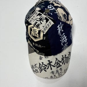 May include: A baseball cap featuring a patchwork design of dark blue, black, and cream fabric panels. The crown displays various Japanese kanji characters and graphic patterns. The off-white brim is printed with black Japanese text and numbers.