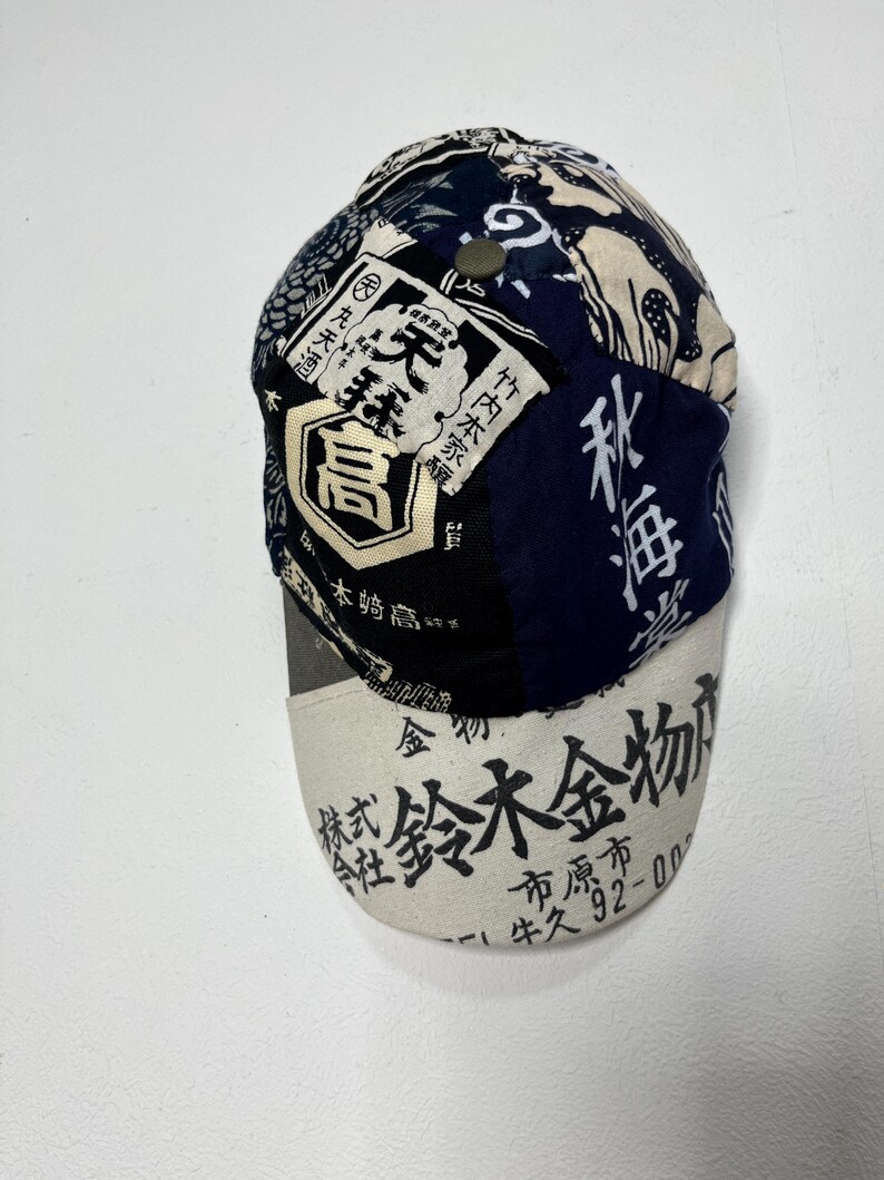 May include: A baseball cap featuring a patchwork design of dark blue, black, and cream fabric panels. The crown displays various Japanese kanji characters and graphic patterns. The off-white brim is printed with black Japanese text and numbers.