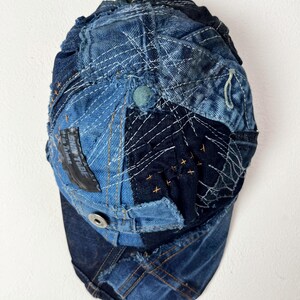 Op de afbeelding: Een blauwe denim baseballpet gemaakt van patchwork stof. De pet heeft verschillende stiksels en een zwart patch met de tekst "Sun Tribe" op de voorkant.