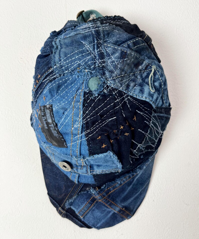Op de afbeelding: Een blauwe denim baseballpet gemaakt van patchwork stof. De pet heeft verschillende stiksels en een zwart patch met de tekst "Sun Tribe" op de voorkant.