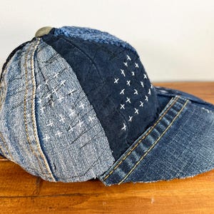 May include: A blue denim baseball cap with a patchwork design. The cap is made of different shades of blue denim and features white stitching in a cross pattern.