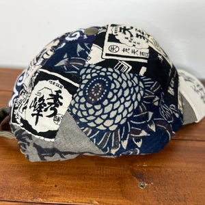 May include: A baseball cap featuring a patchwork design of various fabrics in navy blue, black, white, and grey. The panels display diverse patterns including Japanese kanji characters, floral motifs, and abstract shapes, with a solid button on top.