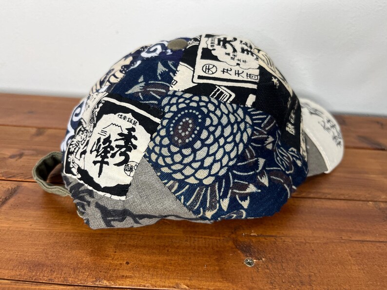 May include: A baseball cap featuring a patchwork design of various fabrics in navy blue, black, white, and grey. The panels display diverse patterns including Japanese kanji characters, floral motifs, and abstract shapes, with a solid button on top.