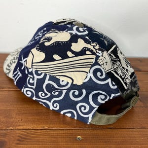 May include: A baseball cap featuring a patchwork design of dark blue and cream fabric with various patterns, including swirling waves and Japanese-style calligraphy. The cap has an olive green adjustable strap at the back with a metal buckle.