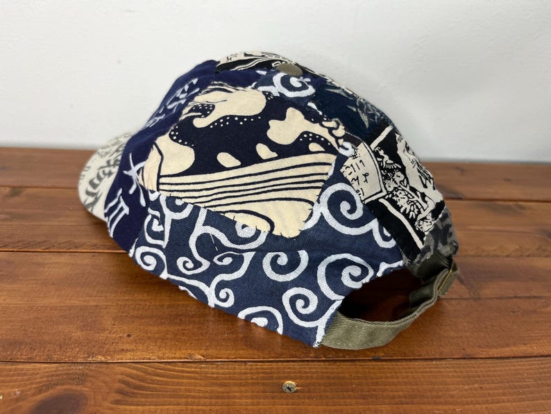 May include: A baseball cap featuring a patchwork design of dark blue and cream fabric with various patterns, including swirling waves and Japanese-style calligraphy. The cap has an olive green adjustable strap at the back with a metal buckle.