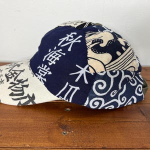 May include: A baseball cap featuring a patchwork design of navy blue and cream fabric. The cap displays Japanese kanji characters, wave patterns, and swirling motifs. The brim is cream-coloured with black text, and the crown is constructed from multiple panels of contrasting fabric patterns.