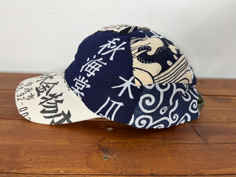 May include: A baseball cap featuring a patchwork design of navy blue and cream fabric. The cap displays Japanese kanji characters, wave patterns, and swirling motifs. The brim is cream-coloured with black text, and the crown is constructed from multiple panels of contrasting fabric patterns.