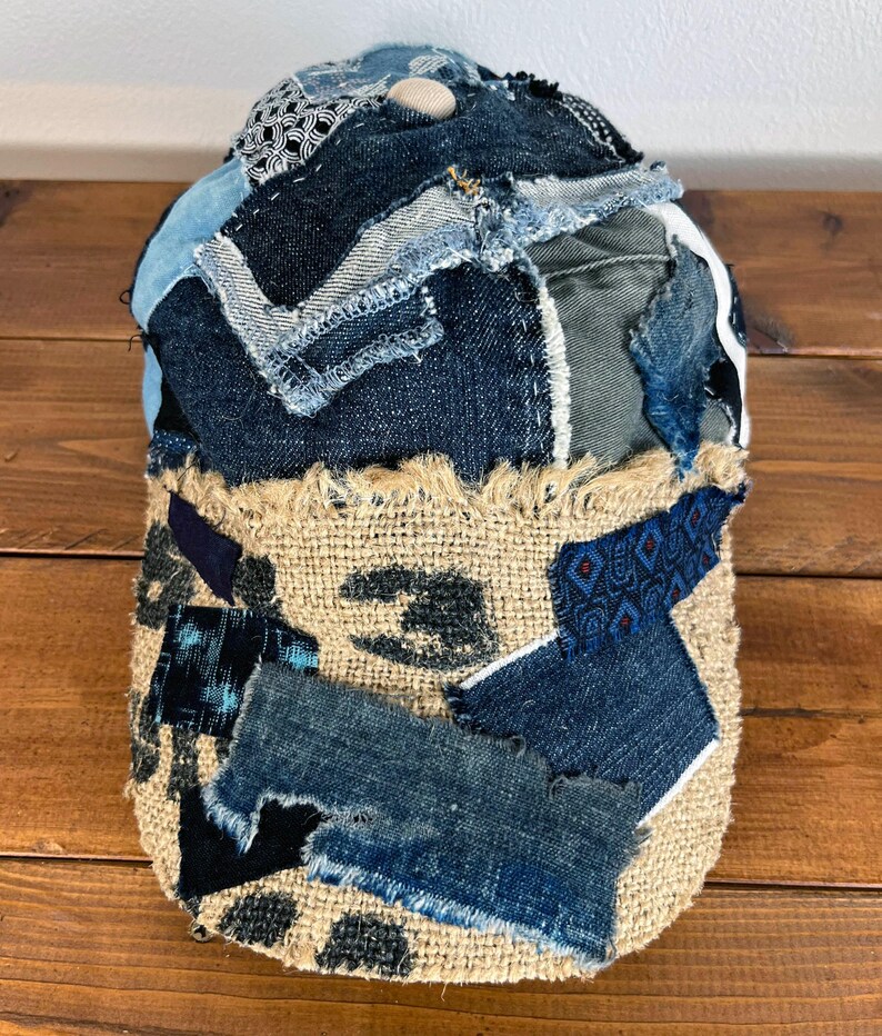Op de afbeelding: Een baseballpet van denim en jute met een patchwork-ontwerp. De pet is gemaakt van verschillende tinten blauw denim en heeft een gescheurde jute rand.