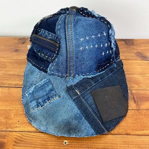 May include: A baseball cap constructed from various shades of blue denim fabric patches. The cap features visible white cross-stitch embroidery, a dark blue zipper detail on the side, frayed edges, and a solid black square patch on the brim.