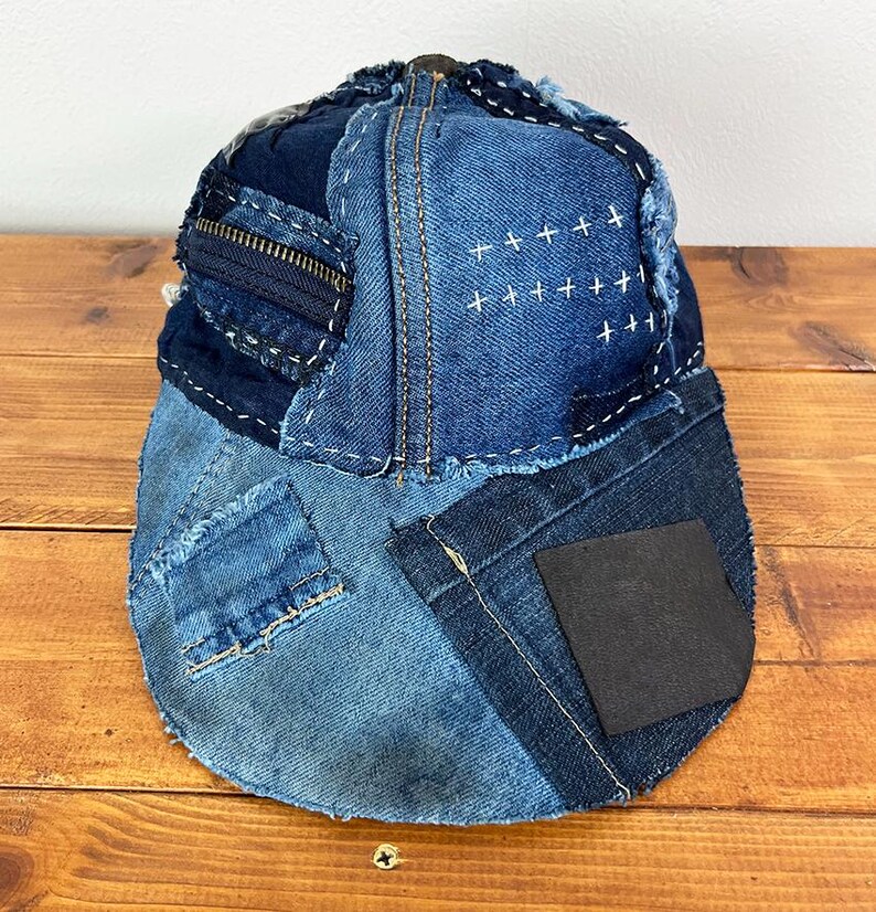 May include: A baseball cap constructed from various shades of blue denim fabric patches. The cap features visible white cross-stitch embroidery, a dark blue zipper detail on the side, frayed edges, and a solid black square patch on the brim.