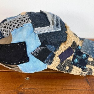 Op de afbeelding: Een bruine, blauwe en zwarte patchwork baseballcap met een gebogen klep. De cap is gemaakt van verschillende stoffen, waaronder denim, jute en katoen.