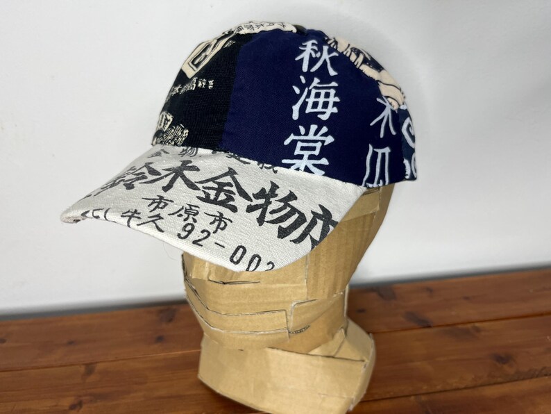 May include: A baseball cap featuring a white brim with black Japanese text and numbers, and a crown composed of black and navy blue panels. The navy blue panel displays vertical white Japanese characters, while the black panel features graphic patterns.