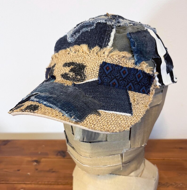 Op de afbeelding: Een bruine en blauwe patchwork baseballcap met een gefranst vizier. De cap is gemaakt van verschillende stoffen, waaronder denim, jute en een stof met een patroon.