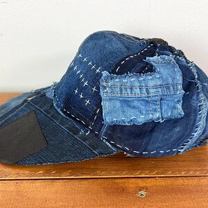 May include: A baseball cap constructed from various shades of blue denim fabric. The cap features visible white stitching, patches of different denim textures, and a dark rectangular accent on the brim, showcasing a layered, pieced-together design.