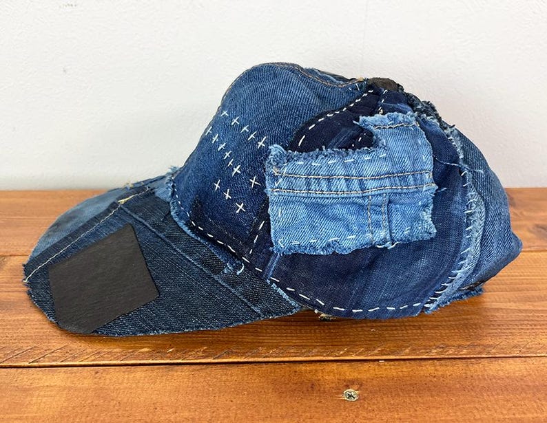 May include: A baseball cap constructed from various shades of blue denim fabric. The cap features visible white stitching, patches of different denim textures, and a dark rectangular accent on the brim, showcasing a layered, pieced-together design.