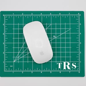 Puede incluir: Un ratón Apple Magic blanco sobre una esterilla de corte verde con líneas de cuadrícula y medidas en centímetros. La esterilla tiene las letras "TRS" en la esquina inferior derecha. El ratón está centrado en la esterilla.