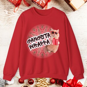 Pode incluir: Camisola vermelha com o texto "Gangsta Wrappa" e um gráfico de um gato usando óculos de sol em forma de coração e segurando um presente. O gato está em um fundo circular com um padrão decorativo.