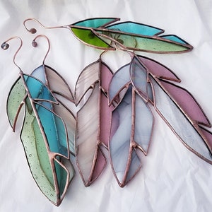 May include: Stained glass leaf ornaments in shades of green, blue, pink, and white, with copper wire detailing. The leaves are arranged in a fan shape, with a curved wire at the top for hanging. The glass has a textured appearance.