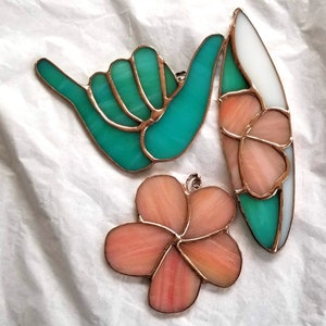 Pode incluir: Tr&ecirc;s ornamentos de vitrais: uma m&atilde;o turquesa no sinal shaka, uma prancha de surf com sec&ccedil;&otilde;es brancas, turquesas e p&ecirc;ssego, e uma flor de p&ecirc;ssego. Cada pe&ccedil;a tem uma borda cor de cobre e uma argola para pendurar.