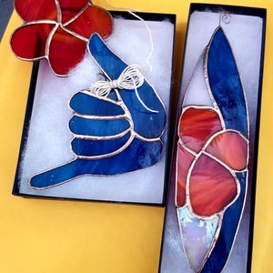 Pode incluir: Tr&ecirc;s ornamentos de vitrais em caixas pretas. Um &eacute; uma flor vermelha, outro &eacute; um gesto de m&atilde;o azul e o terceiro &eacute; uma forma de prancha de surf com se&ccedil;&otilde;es vermelhas, azuis e brancas. Cada um tem uma moldura de cobre e uma corda para pendurar.