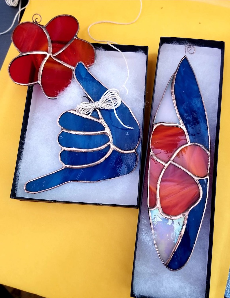 Pode incluir: Tr&ecirc;s ornamentos de vitrais em caixas pretas. Um &eacute; uma flor vermelha, outro &eacute; um gesto de m&atilde;o azul e o terceiro &eacute; uma forma de prancha de surf com se&ccedil;&otilde;es vermelhas, azuis e brancas. Cada um tem uma moldura de cobre e uma corda para pendurar.