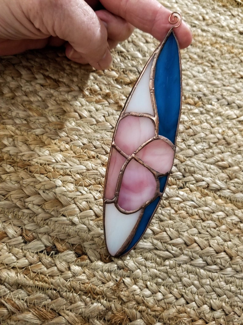 Pode incluir: Um ornamento de vitral oval. Apresenta sec&ccedil;&otilde;es de vidro branco, rosa e azul, contornadas com metal cor de cobre. Uma argola de arame de cobre est&aacute; no topo para pendurar. Mede aproximadamente 15 cm de comprimento.