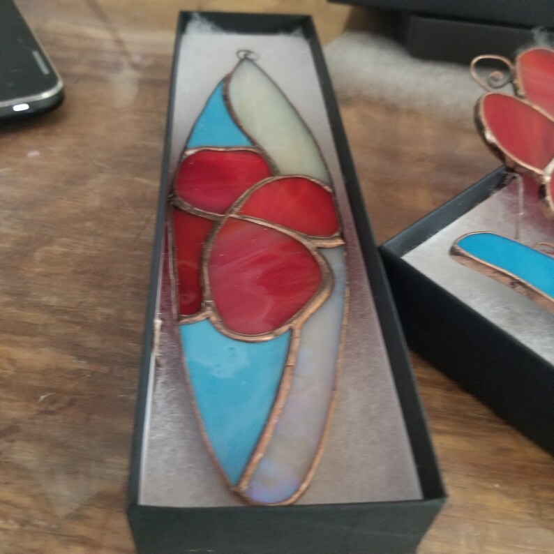 Pode incluir: Um ornamento de vitral numa caixa preta. O ornamento tem formato de gota com pe&ccedil;as de vidro vermelhas, azul claro e brancas, delineadas com cobre. O ornamento &eacute; projetado com formas abstratas e est&aacute; pronto para pendurar.
