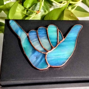 May include: A stained glass shaka hand gesture in shades of blue and turquoise with copper edging. The artwork is displayed on a black box, with green foliage in the background. A decorative piece.