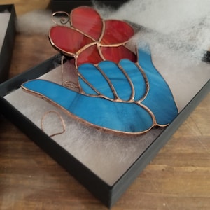 Pode incluir: Um ornamento de vitral numa caixa preta. O ornamento apresenta uma m&atilde;o azul com uma flor vermelha. O vidro &eacute; delineado em cobre. O ornamento est&aacute; sobre material de embalagem branco.