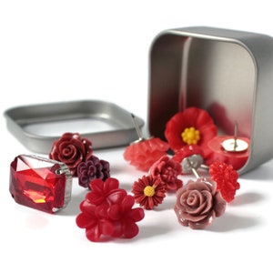 May include: A collection of decorative push pins in various shades of red and pink, including floral and gem designs. Some pins are scattered around an open, square, silver tin. The pins are on a white background.