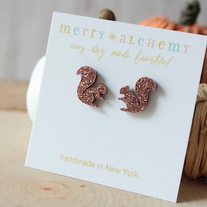 May include: A pair of rose gold glitter squirrel earrings on a white card with the text "merry*alchemy every day made beautiful" and "handmade in New York".