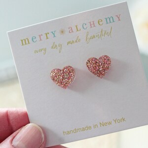 May include: A pair of heart-shaped stud earrings with a glittery pink and gold finish. The earrings are displayed on a white card with the text "merry alchemy every day made beautiful" and "handmade in New York".