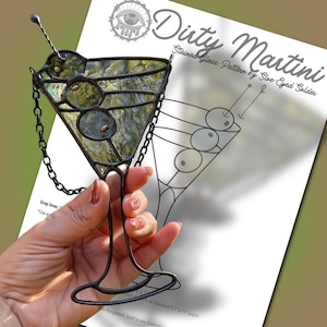 May include: A stained glass martini glass ornament with three olives, featuring dark metal solder lines and a hanging chain. Behind it is a printed paper pattern titled Dirty Martini Stained Glass Pattern by Sloe-Eyed Solder with line drawings.