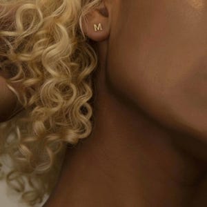 Peut inclure: Gros plan sur une oreille portant une boucle d'oreille en or avec la lettre "M". La personne porte également un délicat collier en or. L'image présente une palette de couleurs chaudes et neutres.