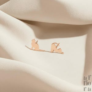 May include: A pair of rose gold cat-shaped stud earrings. One cat is sitting up, and the other is stretching. The earrings are displayed on a cream-colored fabric. The brand name "af fiora" is in the bottom right corner.