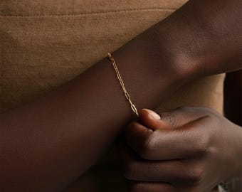 May include: A delicate gold chain bracelet with an elongated link design. The bracelet is worn on a dark-skinned arm, with the hand gently holding the bracelet. The background is a solid, warm brown color.