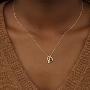 Op de afbeelding: Een delicate gouden ketting met een hanger met de letters "HD" en een druppelvormige groene edelsteen. De ketting wordt gedragen op een bruine trui.