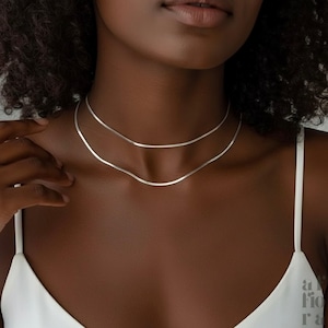 May include: Two delicate, silver necklaces. The necklaces are thin and snake-chain style, resting on the collarbone. The model is wearing a white spaghetti strap top. The background is a neutral colour.