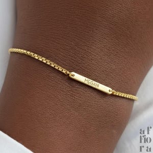 May include: A gold-colored bracelet with a rectangular bar engraved with the name "Megan". The bracelet has a delicate chain design. The image is a close-up shot of the bracelet on a person's wrist.