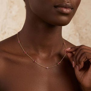 May include: A delicate gold necklace featuring small, round, red gemstones evenly spaced along the chain. The necklace is worn around the neck, with the clasp visible. The gemstones are a vibrant red color, and the chain is a warm gold tone.