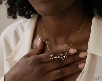 Peut inclure: Un collier doré avec le nom "Emily" écrit en cursive. Le collier est porté autour du cou, le nom reposant sur la poitrine. La personne porte une chemise blanche.